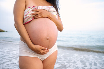 Fototapeta na wymiar Pregnant woman caressing her bare belly with her hands standing on a beach at sunset with a delicate evening light glowing on the horizon over the sea with copyspace
