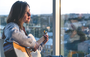 A young woman plays an acoustic guitar by the window.