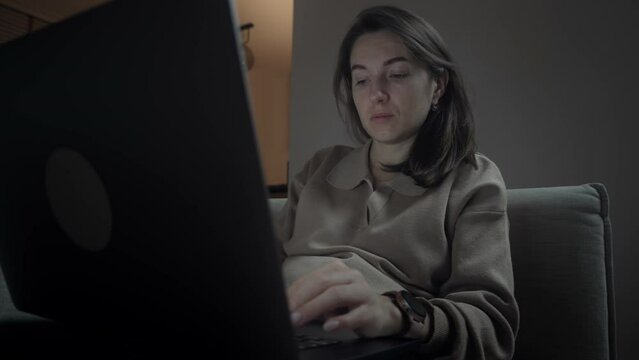 Close-up of concentrated adult woman browsing laptop late in the evening. Serious female worker or freelancer sitting on couch in her living room in front of computer screen at night, concept of