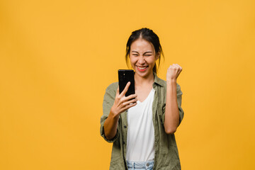 Surprised young Asia lady using mobile phone with positive expression, smiles broadly, dressed in casual clothing and stand isolated on yellow background. Happy adorable glad woman rejoices success.