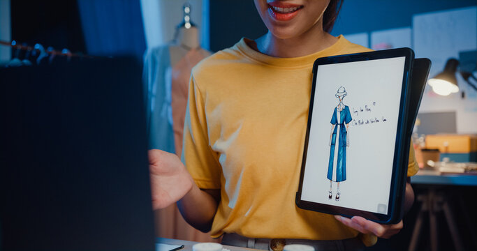 Close-up young Asia lady dressmaker use digital tablet meeting with designer discuss show sample dress design new collection on screen on desk in shop at night. Tailor new business startup concept. - Powered by Adobe