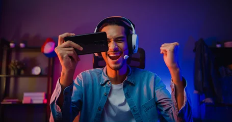 Fotobehang Gaming Happy asia man gamer wear headphone competition play video game online with smartphone colorful neon light in living room at night modern house. Esport streaming game online, Home quarantine activity.  © tirachard