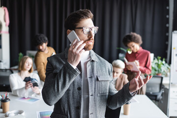 advertising manager in eyeglasses talking on smartphone near blurred multicultural colleagues.