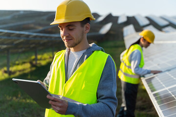 Engineer of solar power plant blogging on company page on social network. Young man in yellow vest holding tablet in hands, posting new content on background of solar panels