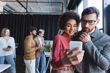 smiling african american woman showing smartphone to thoughtful colleague in ad agency.