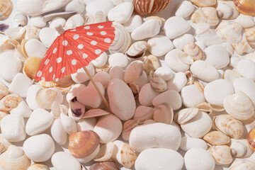 Red sun umbrella with sunlight shadow on different sea shells and white stones. Minimal summer vacation concept. Creative beach relaxation idea.
