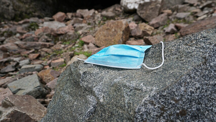 The used medical mask was thrown out in nature. Environmental pollution. An old medical mask lies on a rock in the mountains. A stream runs, grass grows, a forest, an ice grotto. Danger to others.