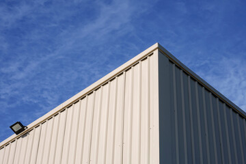 Fototapeta premium Sunlight on surface of white corrugated steel warehouse wall against clouds on blue sky in low angle view