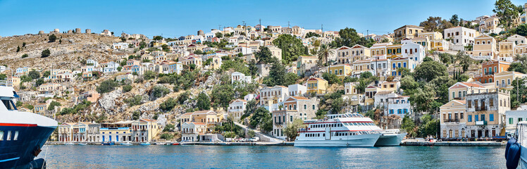Fototapeta premium Multi-colored houses scattered on hills of Symi island view from water. Coastal town on turquoise sea shore in Greece. Tourist liners moor by seafront