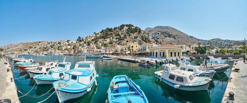 Moored tourist and fishing boats at pier in port of island Symi in Greece. Vessels drift on turquoise water against small multicolored houses on hills