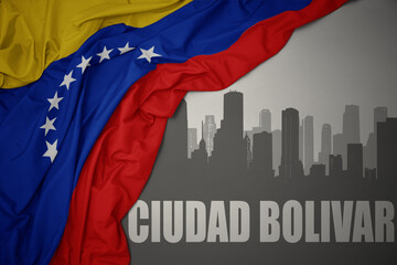 abstract silhouette of the city with text Ciudad Bolivar near waving colorful national flag of venezuela on a gray background.