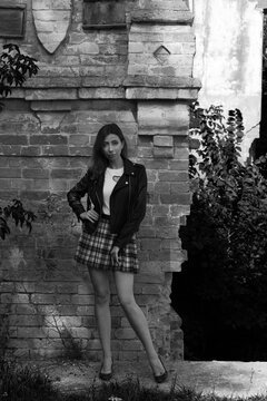 Slender woman in short plaid skirt standing near vintage brick wall of an abandoned old house in black and white