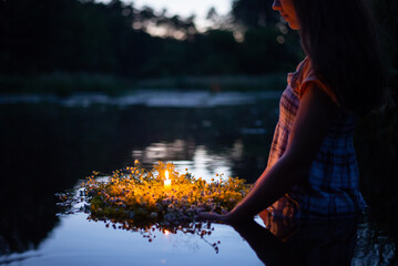 Young girl holding wreath with burning candle, setting it to flow on the river, making a wish - obrazy, fototapety, plakaty