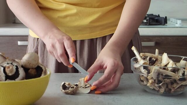 Woman cuts mushrooms with a knife with her hands for soup or sauce. Young woman with a beautiful manicure slices champignons in the kitchen. Close up HD. Brown and yellow colors. Cooks lunch or dinner