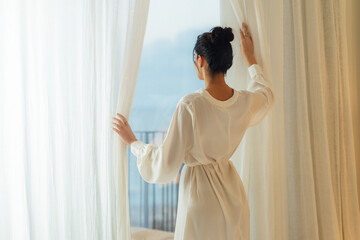 Morning of beautiful young woman opening curtains in bedroom and enjoying of the amazing views....