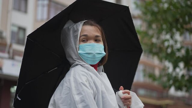 Thoughtful young little woman in coronavirus face mask with umbrella looking away standing outdoors. Portrait of relaxed Caucasian little person resting in city on Covid-19 pandemic