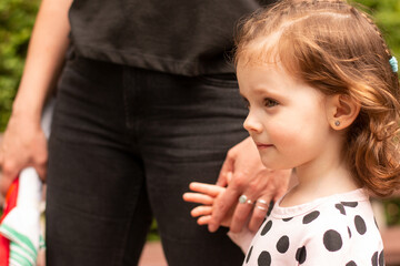 Little beautiful red-haired girl with curly hair in a dress holds the hand of a parent in a summer park