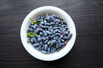 Haskap berry with fresh green leaves in white bowl on wooden table, top view. Honeyberry (Lonicera caerulea)