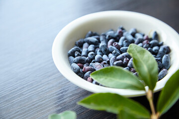 Haskap berry with fresh green leaves in white bowl on wooden table. Honeyberry (Lonicera caerulea)