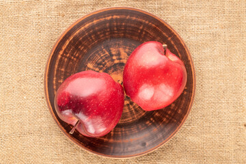 Two ripe red apples in a ceramic plate on burlap, close-up, top view.