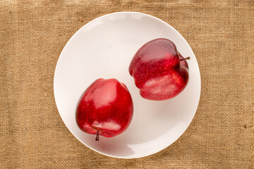 Two ripe red apples in a ceramic plate on burlap, close-up, top view.