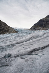 Jostedalsbreen Glacier in Norway flows in a valley between rocky mountain ranges with white, blue, and grey ice forming a rough landscape of cracks and crevasses on a cloudy day. Adventurous hiking. 