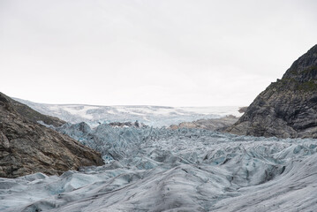 Jostedalsbreen Glacier in Norway forms a rough landscape of cracks and crevasses in the valley between a rocky mountain range. With blue, white, and grey ice. Picture for adventure tour advertisement.