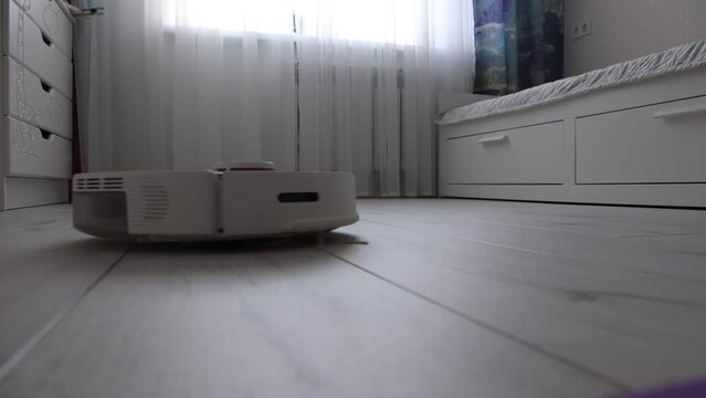 Modern white robot vacuum cleaner vacuums the light floor in the nursery. A smart wireless vacuum cleaner cleans the parquet from dust. Modern automated smart home devices that simplify life.