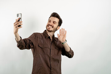 Cheerful young man wearing brown shirt posing isolated over white background taking selfie or having video call and waving hand. Recording vlog, social media influencer streaming, making video call.