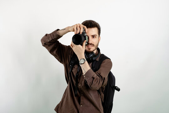 Happy young man wearing shirt posing isolated over white background taking images with dslr camera. Photographer covering his face with the camera.