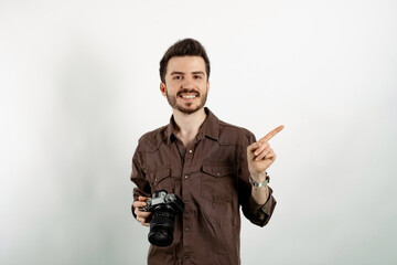 Portrait of young caucasian man wearing shirt posing isolated over white background looking at the camera and pointing index finger aside while holding a professional camera.