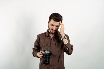 Caucasian man wearing brown shirt posing isolated over white background feeling stressed, unhappy and frustrated, touching forehead and holding dslr camera.