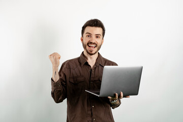 Cheerful caucasian man wearing brown shirt posing isolated over white background using laptop computer while making winner gesture.