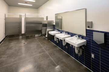 Public School Bathroom with three sinks, mirror, and toilet stalls. No people. Gray and stainless steel. Boys bathroom with urinal and toilet stalls. White Sinks with large mirror
