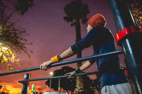 Healthy and fit young latin man with wool cap and wristbands doing calisthenics (resting a moment in bars) on a street workout park with beautiful and colorful sunset sky