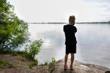 boy traveler stands with his back on a stone and looks pensively at the river