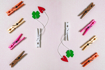 Fototapeta na wymiar Colorful set of different wooden and plastic clothes pegs on a light pink glitter background. A red and a green lucky clover leaf are attached to each of the white wooden clothes pegs. Top view.