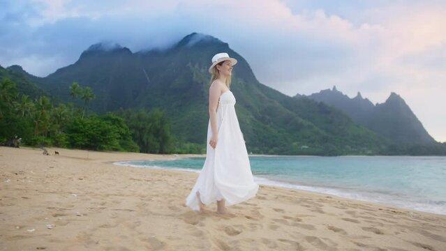 Tropical beach landscape with dramatic grey clouds on top of high green mountains background. Slow motion attractive young woman in trendy boho beach long wedding dress happily walking spinning around