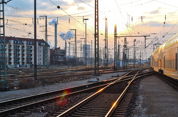 Naklejka na ściany i meble Viele Gleise an einem Bahnhof mit Oberleitungen, Masten, Technik, Signalanlagen, Rauch, Gebäuden und einem Sonnenuntergang