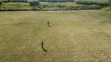 Drone view of a soccer field with two children playing, without identifying the face, and in the background the goalposts with a ball.
