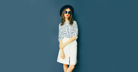 Portrait of beautiful young woman wearing white striped shirt, black round hat on dark blue background