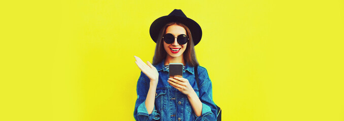 Portrait of stylish happy smiling young woman model with smartphone wearing black round hat, jean jacket on yellow background, blank copy space for advertising text