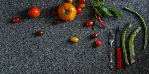 Food border with green asparagus, red and orange tomatoes and red chilli pepper with leaves. Vegetables, red knife and fork on dark grey background  top view, place for text, border