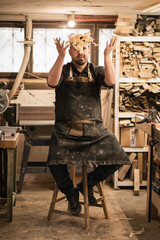 Vertical playful male carpenter throwing up wooden shaving chips in the air sitting on chair, finishing day in workshop