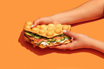 Hand holding bubble waffle sandwich with salmon, arugula, lettuce, cream cheese and cucumber  isolated on orange background. Fast food, take away. Snack