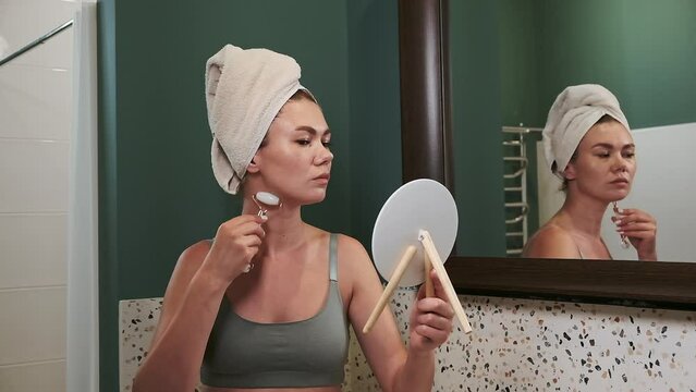 Young woman using jade facial roller for face and neck massage sitting by the bathroom mirror, slow motion