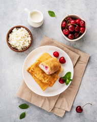 Delicious homemade pancakes or crepes  with cottage cheese and fresh cherries on a white plate with sour cream on a blue background. Top view