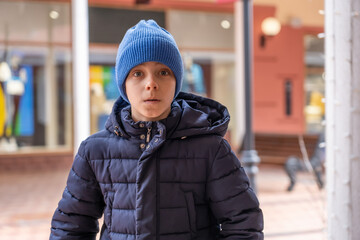 Shocked boy looks into camera and sees staggering prices for fashionable clothes in shop window. Surprised teenager in warm clothes and blue hat looks wide-eyed at camera