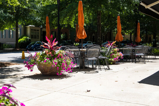 An Outdoor Dinning Area With Black Metal Chairs And Tables And Orange Umbrellas Surrounded By Large Round Flower Pots With Colorful Flowers And Lush Green Trees At Mill Creek Pond In Alpharetta