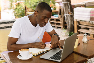 Serious, concentrated and enthusiastic multiracial man using laptop, writing notes in workbook and...